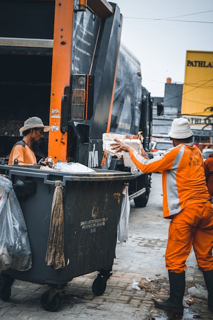 Two waste disposal workers dressed in bright orange high-visibility uniforms and white hats are engaged in a home relocation process involving packing and loading waste materials. One worker is holdin