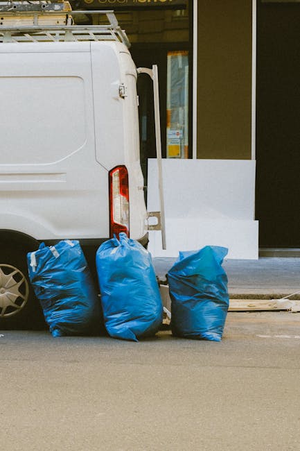 Two waste disposal workers dressed in bright orange high-visibility uniforms and white hats are engaged in a home relocation process involving packing and loading waste materials. One worker is holdin