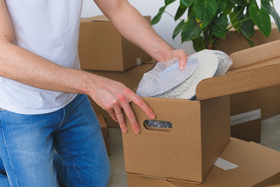 A person wearing a white t-shirt and blue jeans is in a room, carefully removing a fragile antique ceramic dish wrapped in bubble wrap from a brown cardboard box with built-in hand cutouts, part of a 