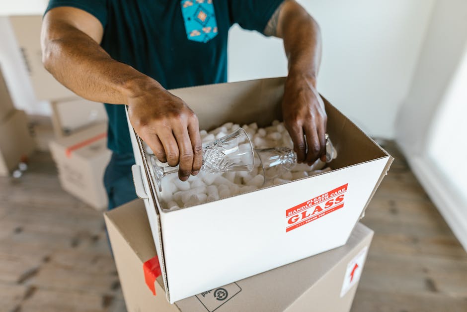 A person wearing a white t-shirt and blue jeans is in a room, carefully removing a fragile antique ceramic dish wrapped in bubble wrap from a brown cardboard box with built-in hand cutouts, part of a 