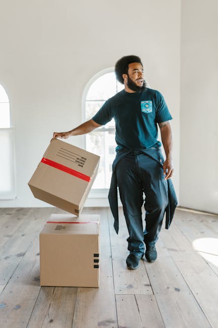 A person dressed in light-colored clothing is stacking cardboard boxes in a room with natural light coming from a large window. The boxes vary in size and are placed on a wooden floor, with some alrea