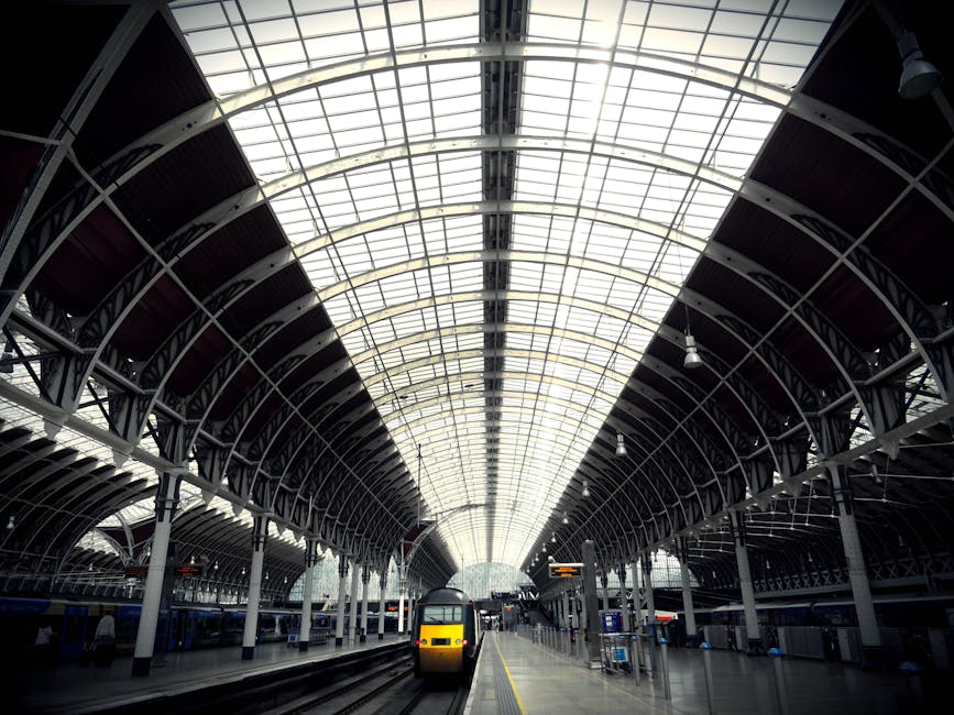 Inside a large train station with a high, arched ceiling made of steel and glass, a modern yellow and black train is stationary on the platform. The platform area shows a row of upright metal barriers