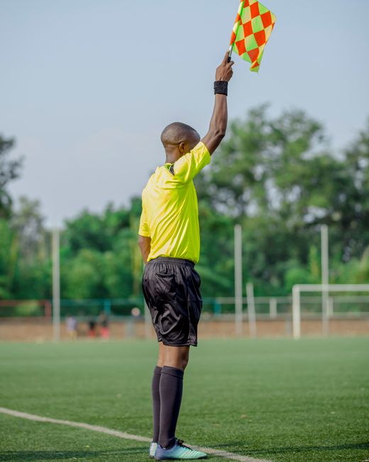 A male referee stands on a grassy sports field during a game, wearing a bright yellow shirt, black shorts, and black knee-high socks. He is holding a brightly patterned, orange and yellow checkered flag high above his head, signaling a decision. The referee has a shaved head and is facing away from the camera. In the background, there are trees and sports equipment, with a clear sky overhead. The setting appears to be an outdoor soccer or football match, with a focus on the referee's role in officiating the game. Paddington Removals, which offers professional removals and house relocation services, operates in environments similar to this outdoor scene, where careful handling and transport of items are essential during home moves. This image emphasizes the importance of clear communication and precise actions, akin to the loading, packing, and transport processes involved in furniture transport and home relocation, within the context of complying with local regulations like the skip and waste rules in Paddington.