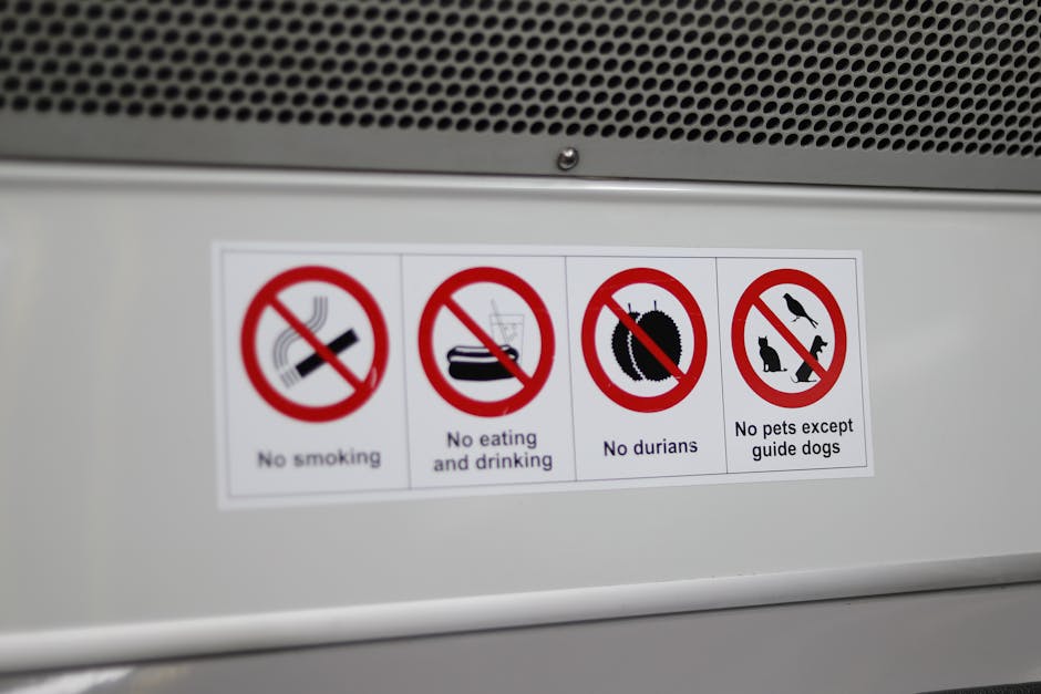 A rectangular informational sign affixed to the exterior of a silver metal vehicle, likely a move or delivery van, displaying four safety and conduct restrictions relevant to house removals and packing processes. The sign shows four separate symbols with red circles and diagonal lines indicating prohibitions: a lit cigarette for no smoking, a food container with a straw for no eating and drinking, a durian fruit for no durians, and a group of pets including a cat, a dog, and a bird for no pets except guide dogs. The sign is mounted just below a perforated metal panel that covers part of the vehicle's body, with natural daylight illuminating the scene. The setting suggests a loading or unloading context within a home relocation operation involving furniture, boxes, and packing materials within a move scenario, with visual cues supporting move logistics and transport safety protocols. Paddington Removals occasionally handles such loading and sorting tasks as part of their professional house removals service.