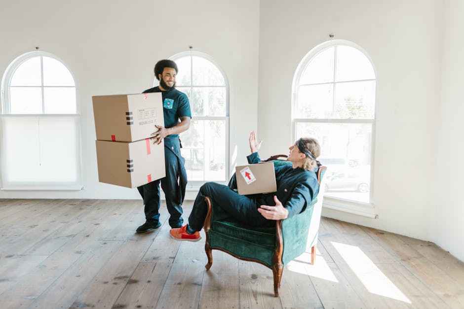 A professional removals technician from Paddington Removals is inside a bright, minimally furnished room with large arched windows, loading two cardboard boxes onto a vintage armchair that a seated wo