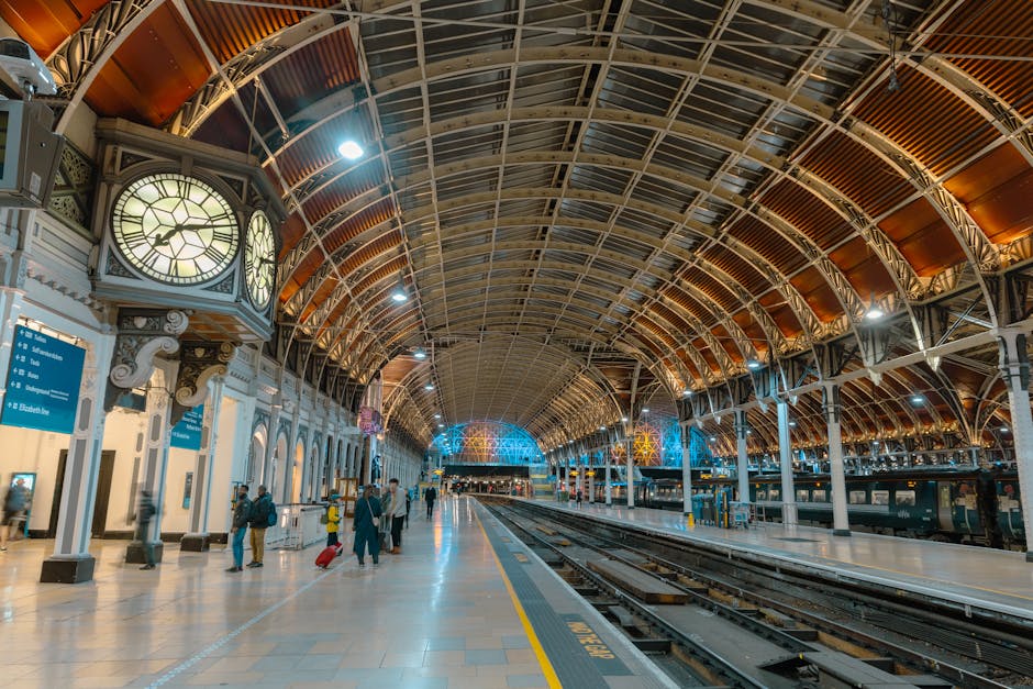 The image depicts a spacious, well-lit train station with an arched, metal and glass ceiling supported by decorative white columns. Inside the station, a few travelers are seen walking along the platf