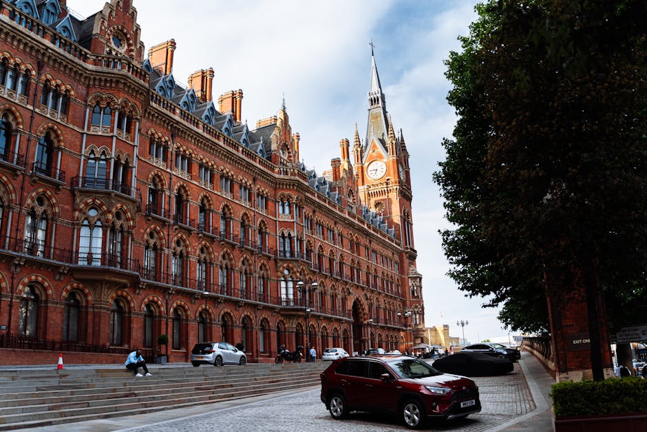 A large red-brick Victorian-style building with ornate architectural details, numerous arched windows, decorative iron balconies, and a tall clock tower featuring a pointed spire and clock face. The b